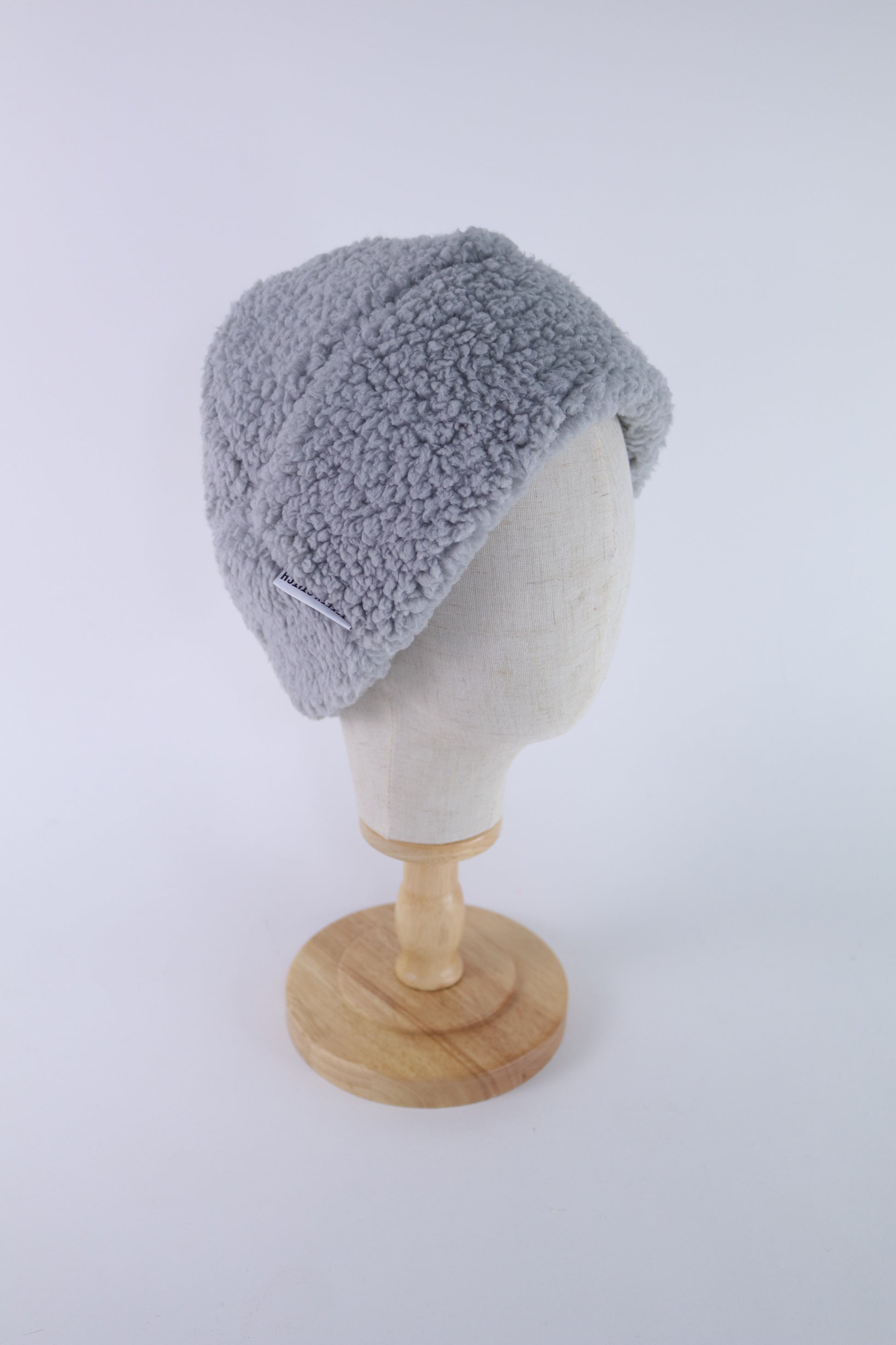Gray faux sherpa beanie displayed on a cream mannequin head with a wooden stand, shown in side view.